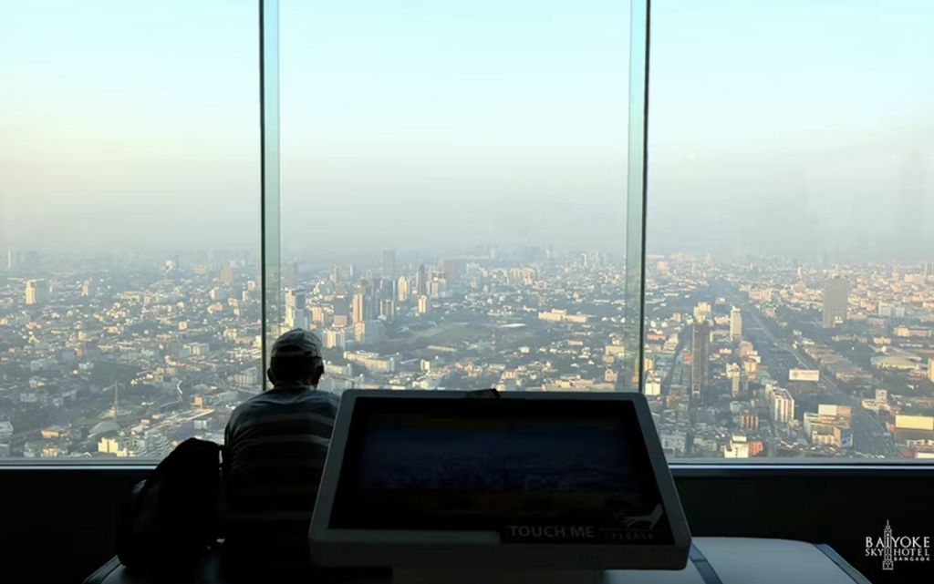 한 사람이 전경에 터치스크린 키오스크가 있는 커다란 유리창을 통해 낮에 펼쳐지는 도시 풍경을 감상하고 있습니다.