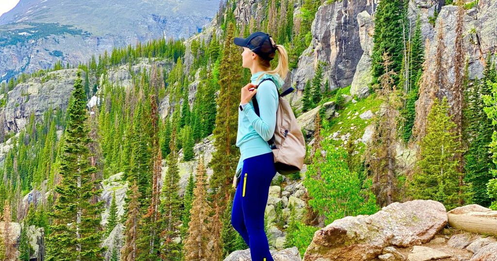 밝은 파란색 상의와 배낭이 달린 남색 바지를 입은 사람이 숲이 우거진 산악 지형의 바위 위에 서 있습니다.