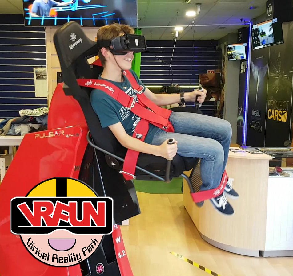 빨간색 하네스가 있는 시뮬레이터 좌석에서 VR 헤드셋을 사용하는 사람, 왼쪽 하단에 "VR FUN Virtual Reality Park" 로고가 있습니다.