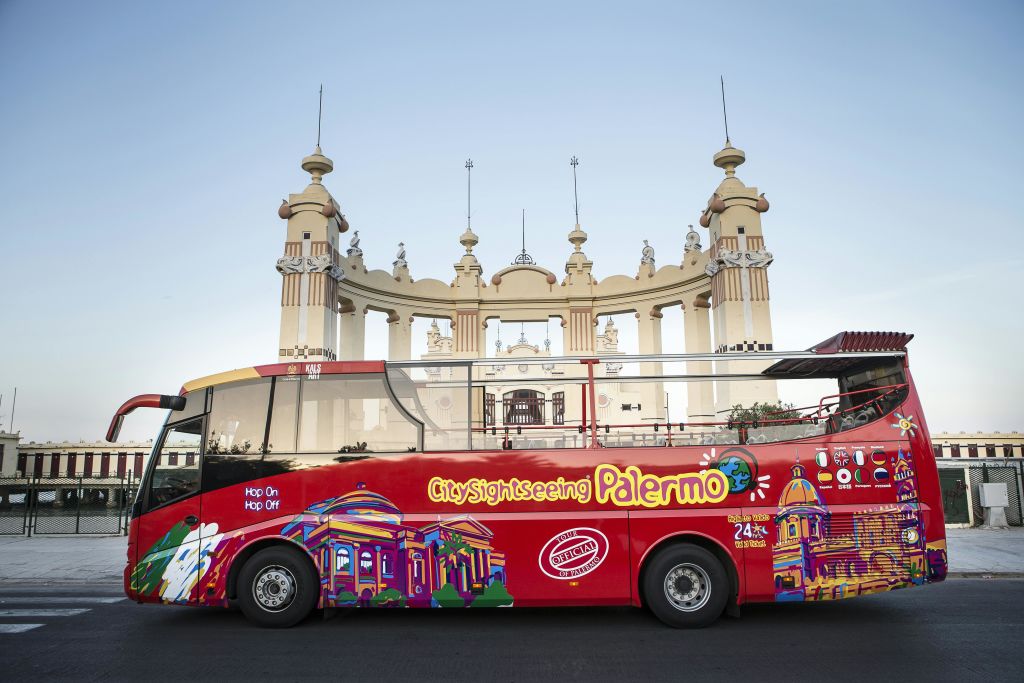 여러 개의 타워가 있는 화려한 유서 깊은 건물 앞에 '팔레르모' 라는 이름의 다채로운 도시 관광 버스가 주차되어 있습니다.
