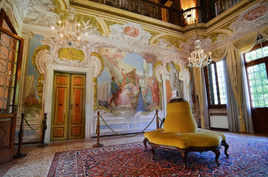 프레스코화로 마감된 벽, 샹들리에, 노란색으로 덮인 벤치, 무늬가 있는 러그가 있는 화려한 객실입니다. 중앙에는 동상이 서 있습니다.