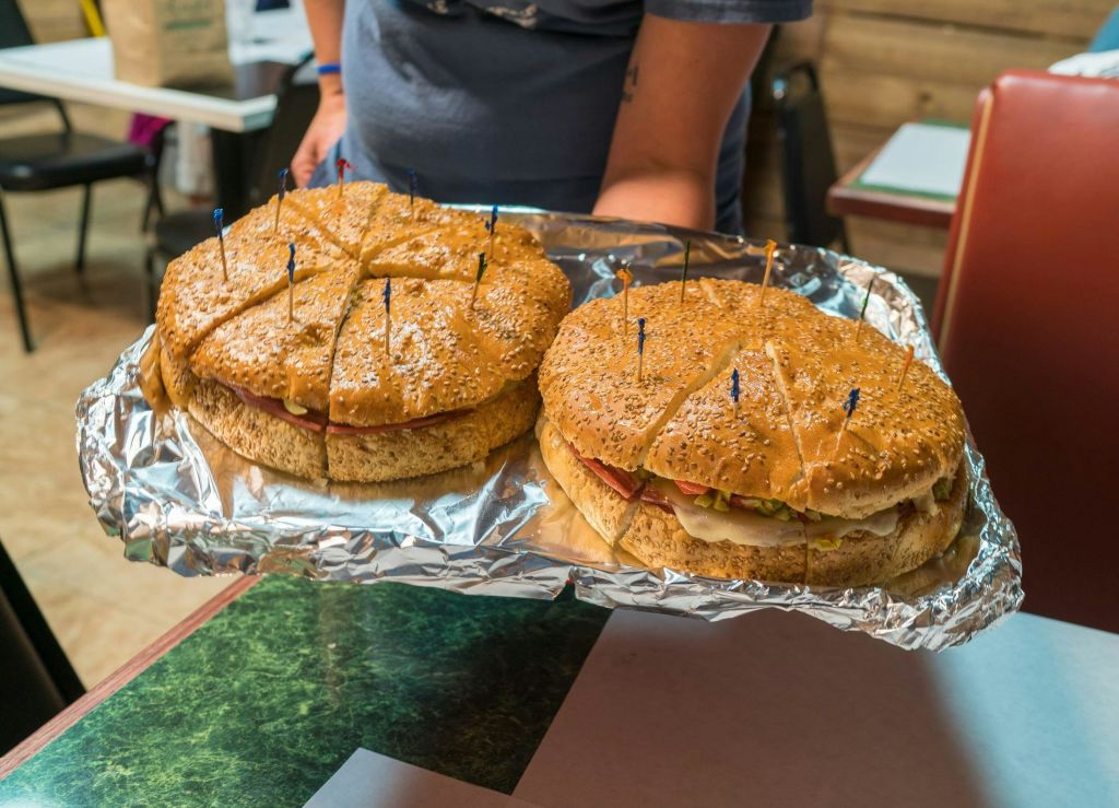 한 사람이 두 개의 커다란 샌드위치가 담긴 트레이를 들고 있으며, 각 샌드위치에는 참깨 만두를 얹고 색색의 이쑤시개로 고정합니다.