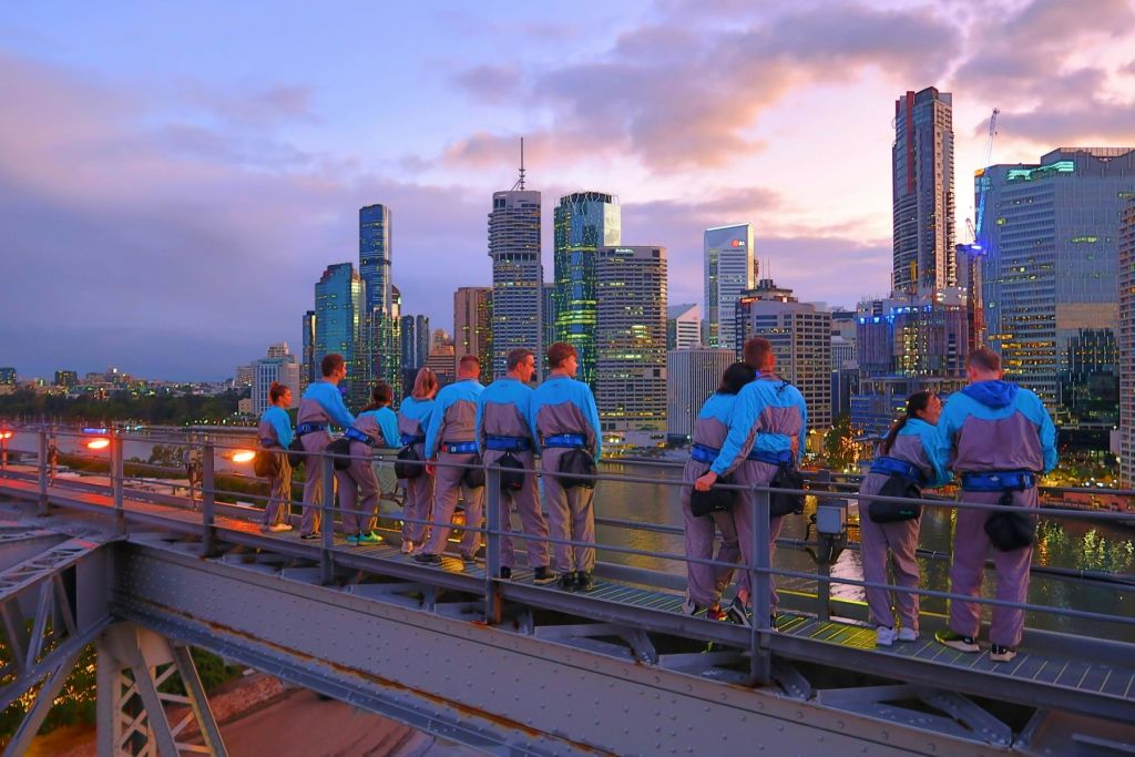 푸른색과 회색 기어를 입은 한 무리의 사람들이 다리에 서서 석양이 질 무렵 도시의 스카이라인을 내려다보고 있습니다.