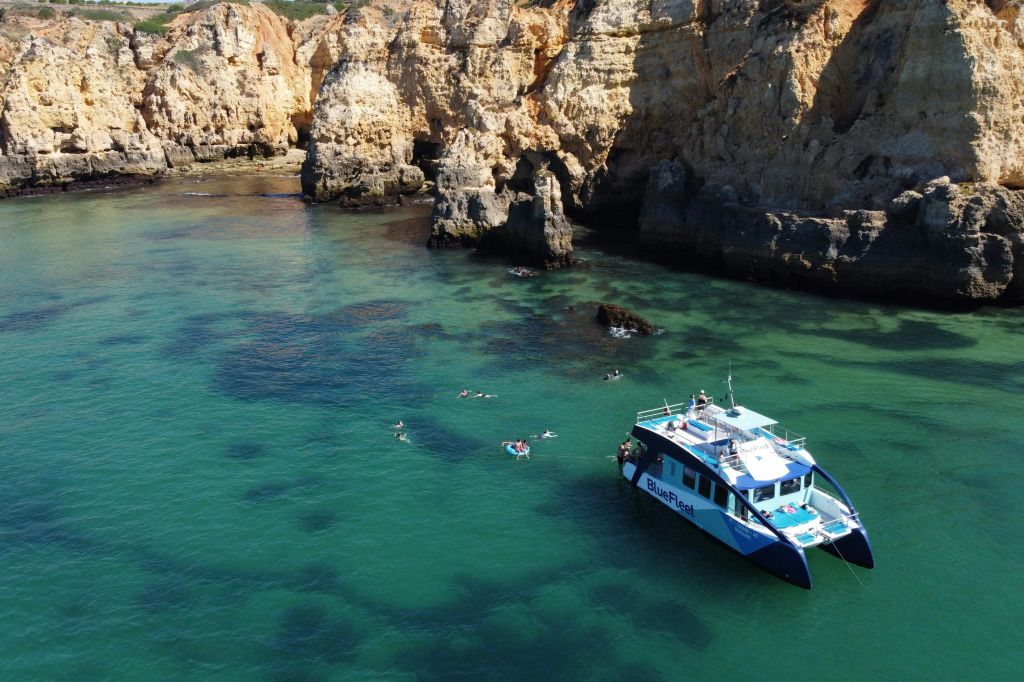 "BlueFleet" 브랜드가 새겨진 쌍동선이 바위 절벽 근처의 맑고 푸른 물에 떠 있습니다. 사람들은 근처에서 수영을 합니다.