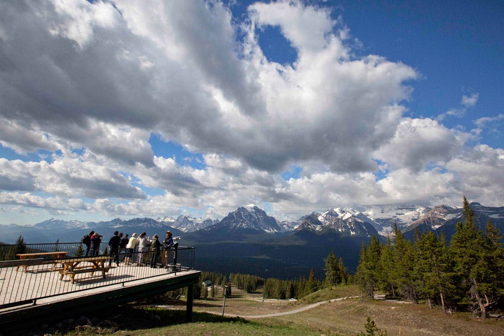 사람들은 흐린 하늘 아래 눈 덮인 산이 내려다보이는 전망대에 서 있으며, 그 아래에는 숲과 길이 있습니다.