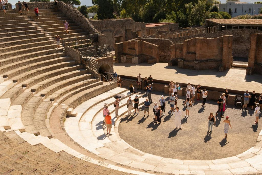 관광객들이 유적과 녹지로 둘러싸인 화창한 하늘 아래 층층이 쌓인 석조 좌석이 있는 고대 원형극장을 탐험합니다.