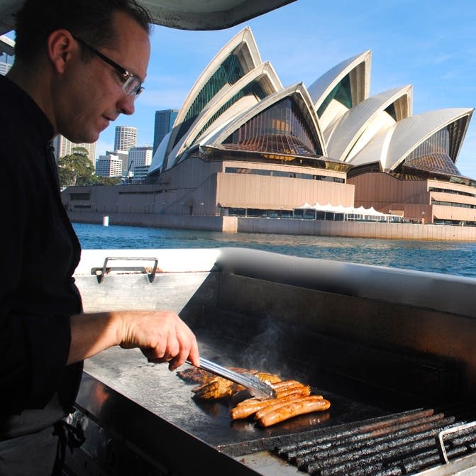 화창한 날 시드니 오페라하우스를 배경으로 한 남성이 바비큐 그릴에서 소시지를 구워줍니다.