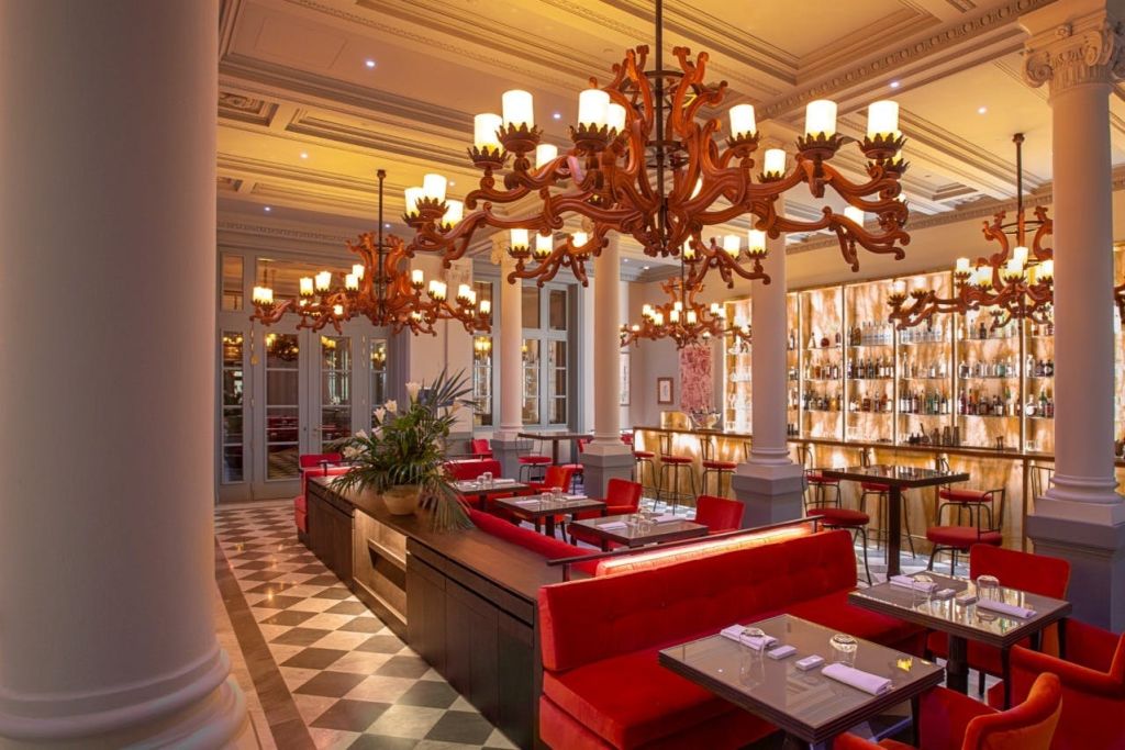 레드 벨벳 부스, 화려한 샹들리에, 체커보드 바닥이 있는 우아한 레스토랑입니다. 병이 표시된 바가 배경에 있습니다.