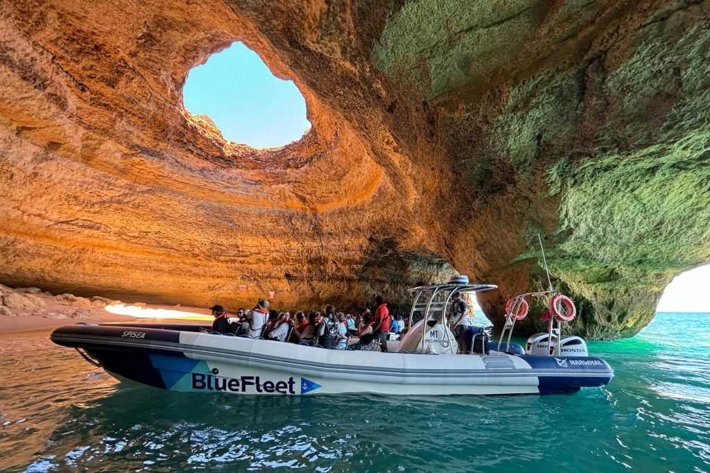 커다란 원형 개구부가 햇빛을 쬐는 바위 동굴 안의 보트에 탄 한 무리의 사람들.