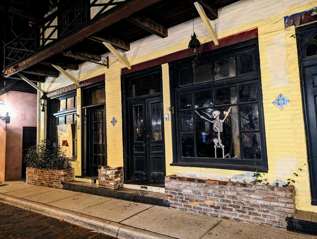 검은색 문과 창문이 있는 노란색 벽돌 건물의 거리 풍경. 창문 중 하나에 해골 장식이 보입니다.