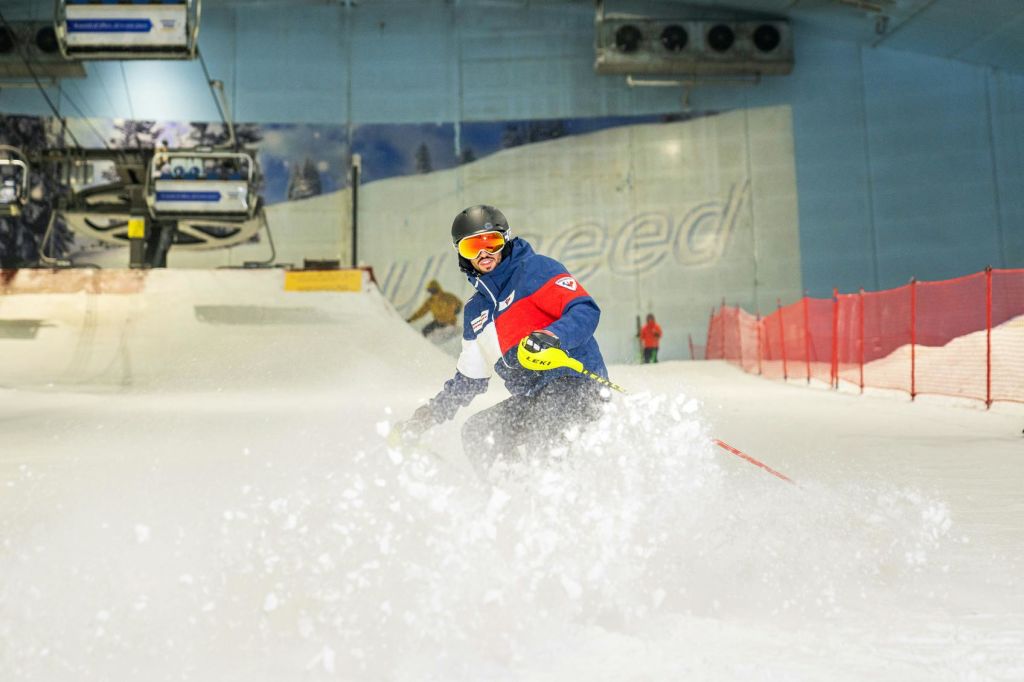 헬멧과 고글을 착용한 스키어가 실내 스키 슬로프에서 자신 있게 미끄러지며 눈을 뿌립니다.