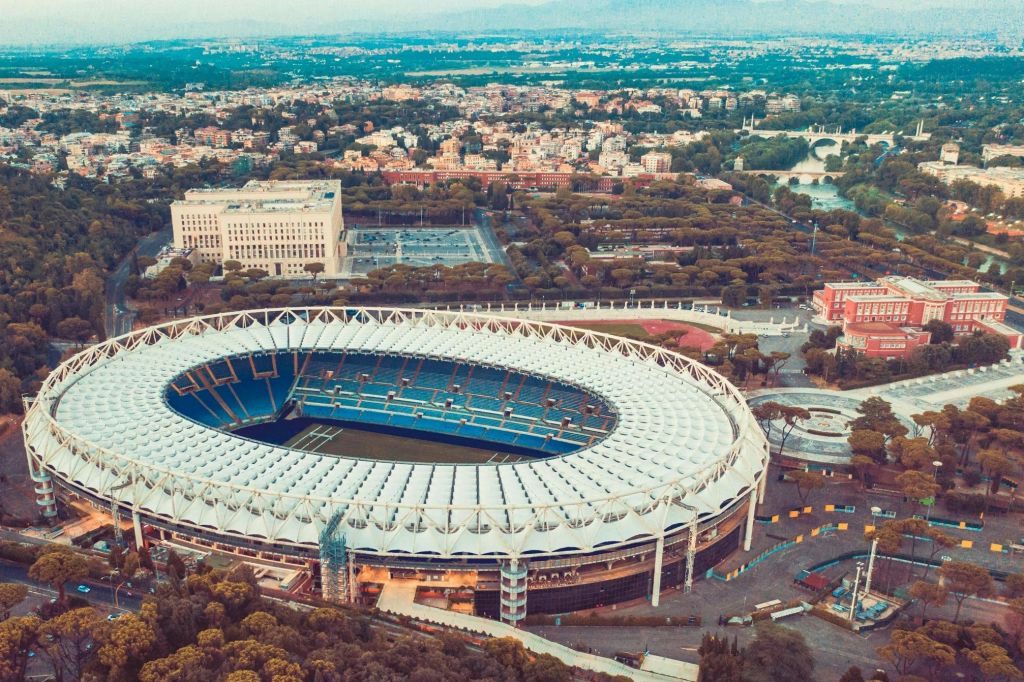 도시 건물, 녹지, 먼 산으로 둘러싸인 하얀 지붕이 있는 대형 경기장의 공중 전망.