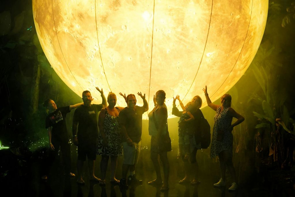 조명이 켜진 커다란 달 모형 앞에 7명이 서서 희미하게 빛나는 환경에서 장난스럽게 포즈를 취하고 있습니다.