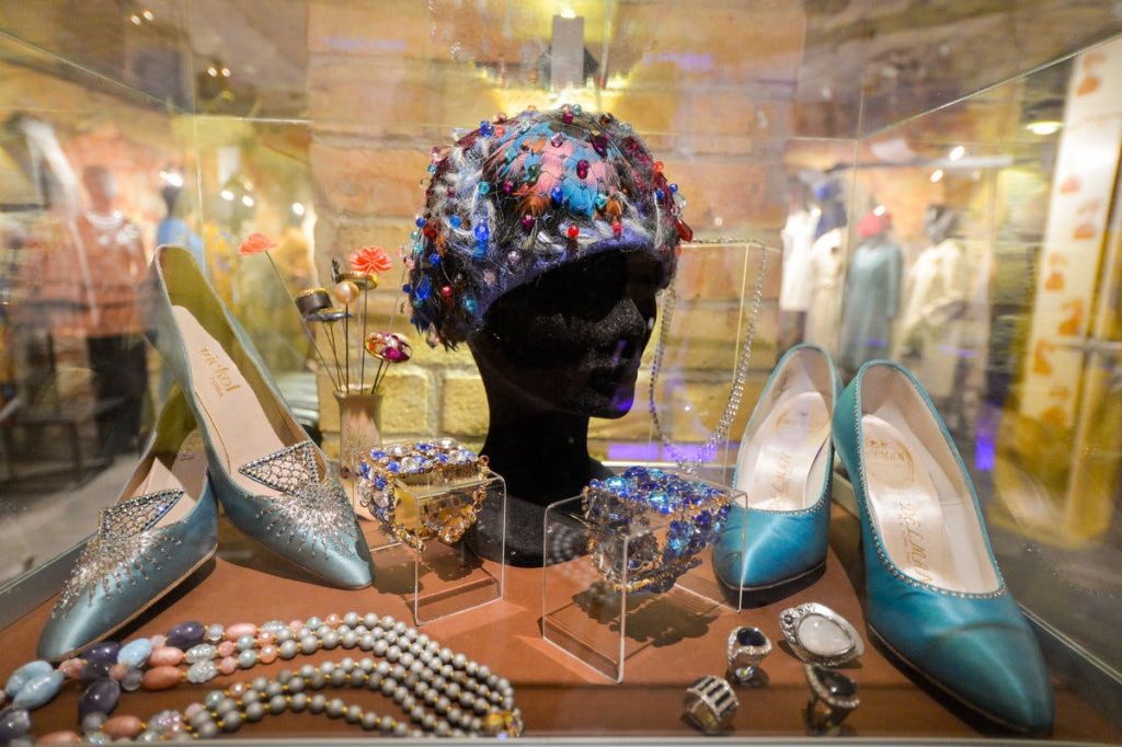 벽돌 배경이 있는 유리 케이스에 구슬 헤드피스, 청록색 신발, 진주 목걸이, 팔찌, 반지를 전시합니다.