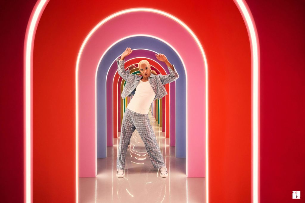 빨간색과 분홍색으로 빛나는 아치가 있는 활기찬 복도에서 무늬가 있는 옷과 운동화를 신고 포즈를 취하고 있는 사람.