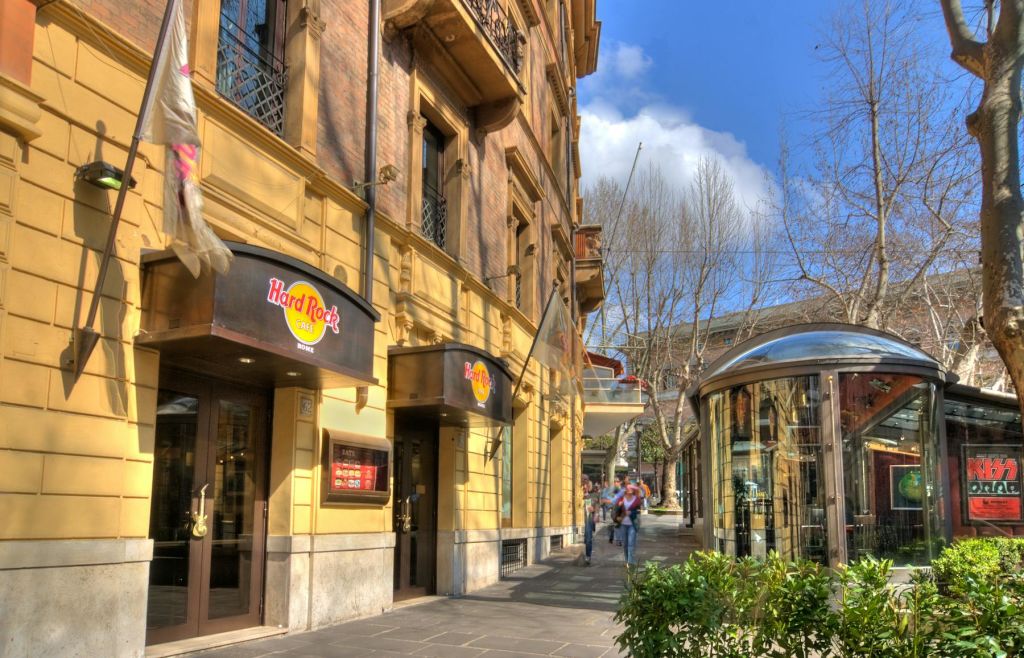 베이지색 유서 깊은 건물에 위치한 하드록 카페의 거리 풍경. 산책하는 사람들과 보도를 가로지르는 나무가 있습니다.