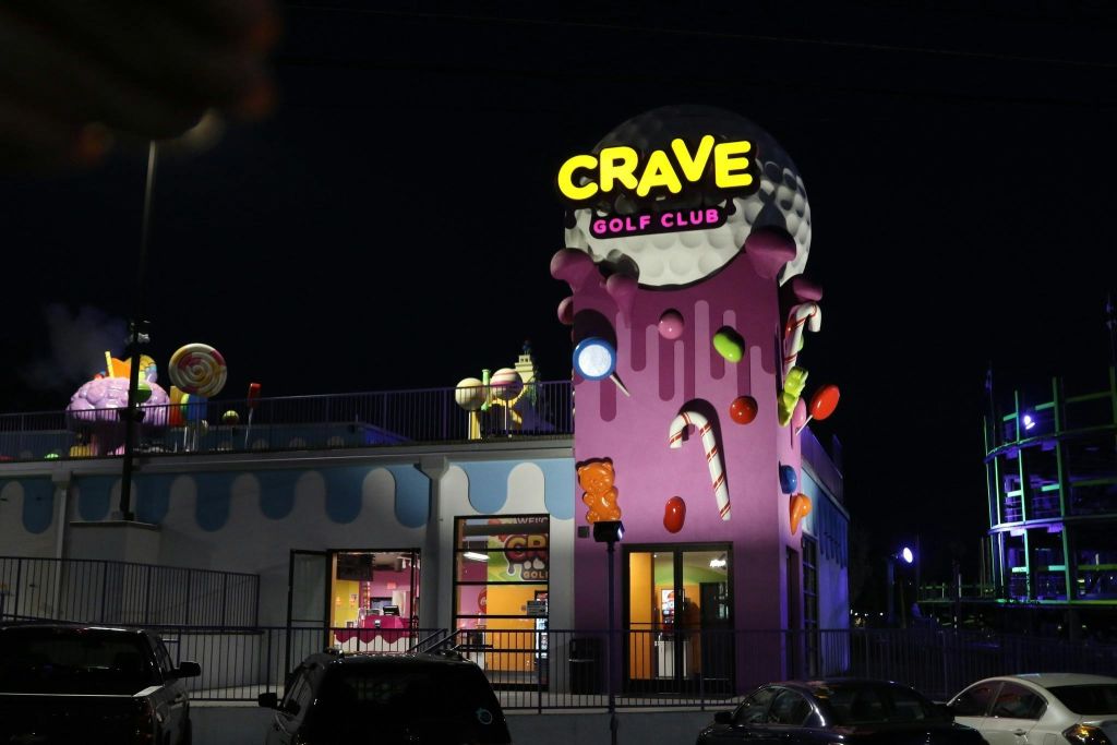 사탕 모양과 아이스크림 테마로 장식된 "CRAVE GOLF CLUB" 간판이 있는 야간 조명이 밝은 건물입니다.