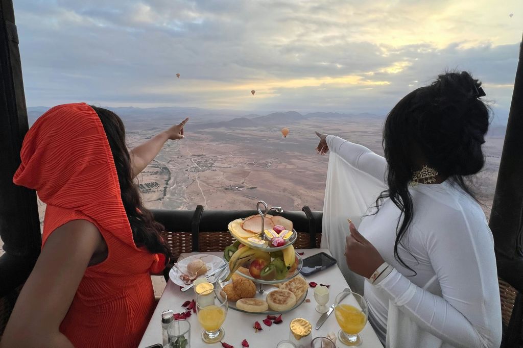 사막 풍경 너머 저 멀리 보이는 다른 열기구들을 가리키며 열기구에서 식사를 즐기는 두 사람.