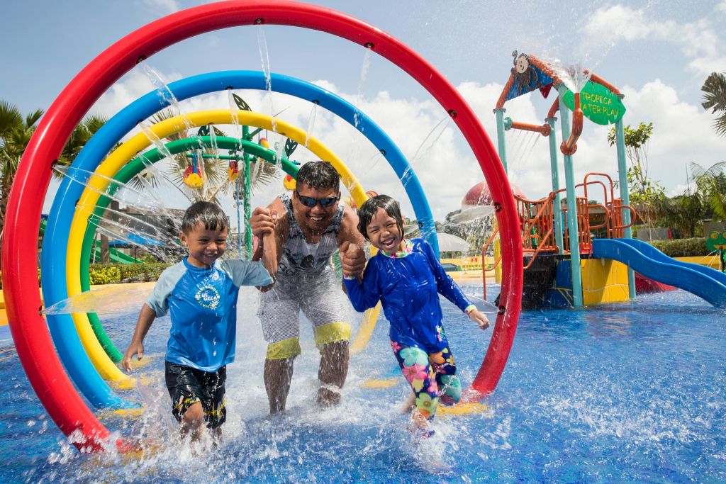 수영복을 입고 미소를 짓는 한 남자와 두 어린이가 워터파크의 다채로운 농구공 아래에서 워터젯을 타고 달리고 있습니다.