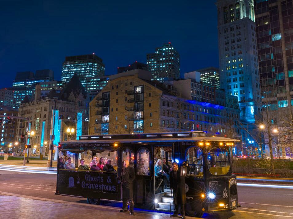 "유령과 묘지" 투어 트롤리가 주차되어 있고, 사람들이 탑승하고, 조명이 켜진 건물을 배경으로 야간 도시 풍경이 펼쳐집니다.