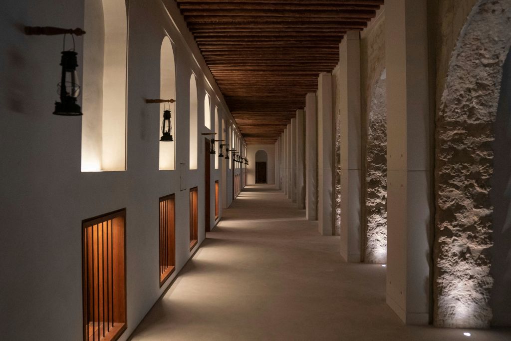 높은 아치, 나무 천장 기둥, 벽에 걸린 작은 등불이 있는 길고 희미한 복도.