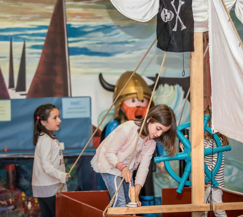 두 소녀가 해적선 테마 놀이 공간에서 놀고 있는데, 한 소녀는 밧줄을 당기고 다른 소녀는 관찰합니다. 배경에는 바이킹 동상이 있습니다.