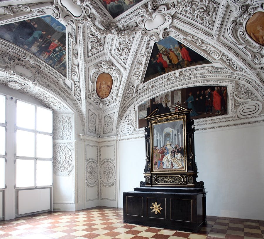 디테일한 천장 프레스코화, 체크무늬 바닥, 커다란 창문, 그림이 있는 중앙의 검은색과 금색 제단이 있는 화려한 객실입니다.