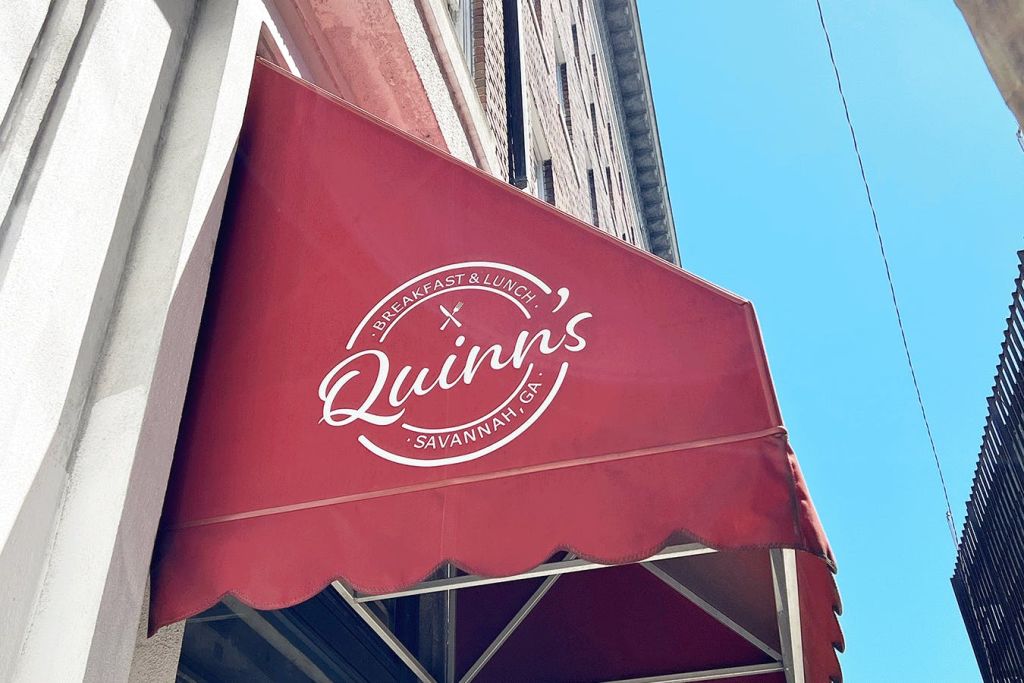 푸른 하늘 아래 벽돌 건물 옆에 흰색으로 'Quinn's Breakfast & Lunch Savannah, GA' 라는 문자가 적힌 붉은 천막.
