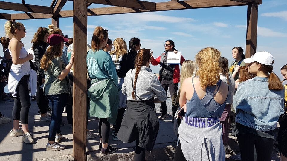 한 무리의 사람들이 종이를 들고 있는 사람 주위에 모여 야외에서 강연이나 발표를 하는 것처럼 보입니다.