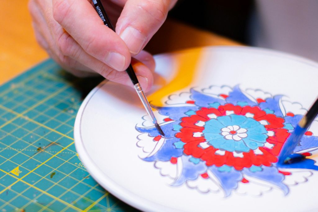 섬세한 브러시를 사용하여 정교한 빨간색, 파란색 및 흰색 꽃 무늬가 있는 흰색 접시를 그리는 손.