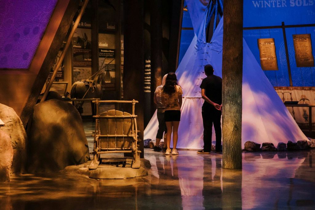 나무로 만든 크래들보드와 정보 디스플레이를 배경으로 조명이 희미한 흰색 티피 근처에 두 사람이 서 있습니다.