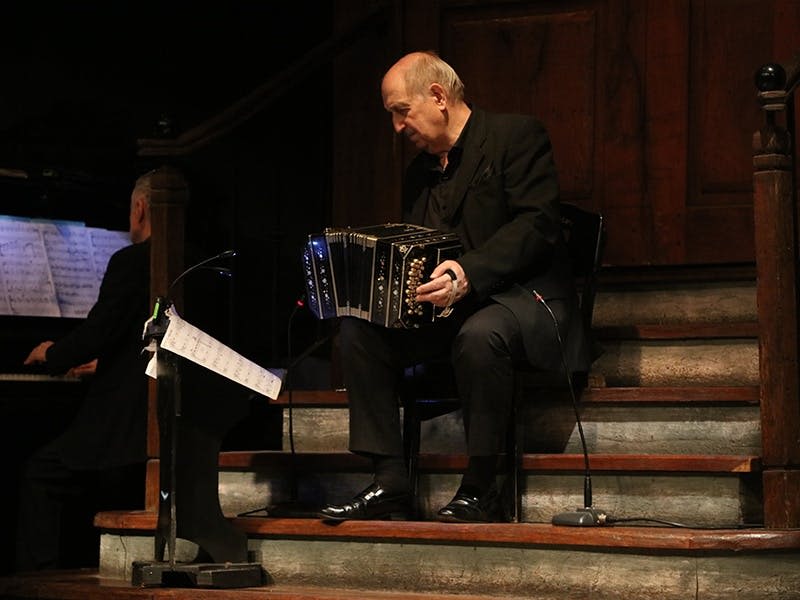 검은 양복을 입은 남성이 나무 계단에 앉아 반도네온을 연주하고 있습니다. 근처에는 악보를 수납할 수 있는 스탠드가 있습니다.