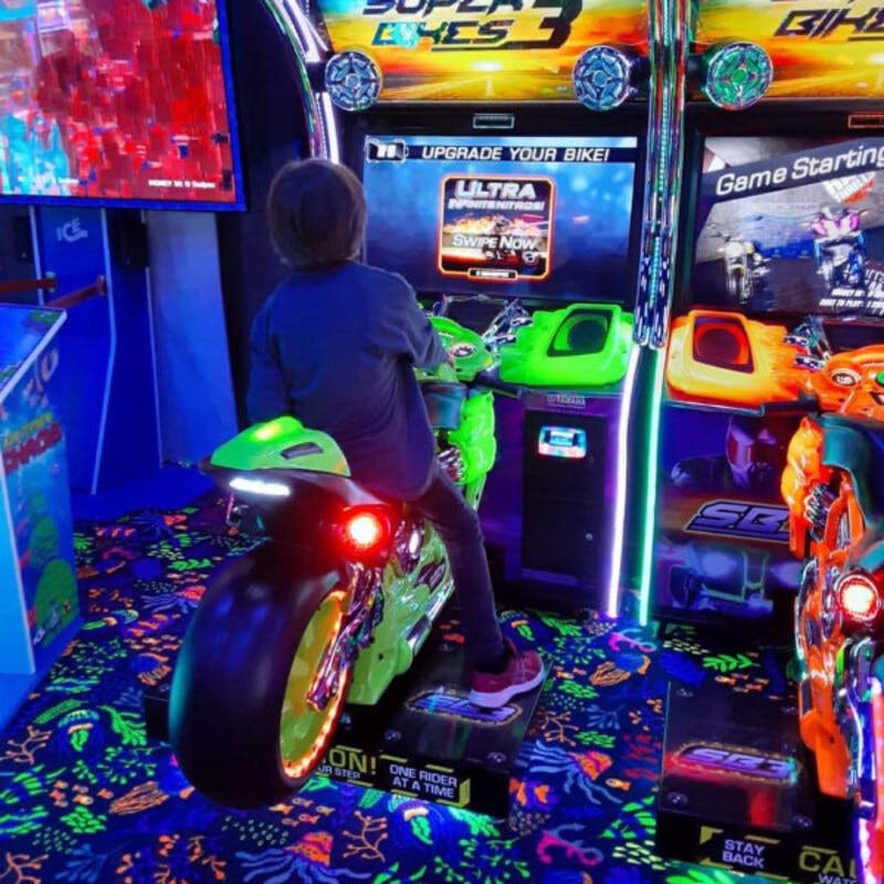다채로운 조명과 그래픽이 돋보이는 녹색 오토바이 아케이드 게임을 타고 네온 패턴의 카펫 위에 서 있는 어린이.