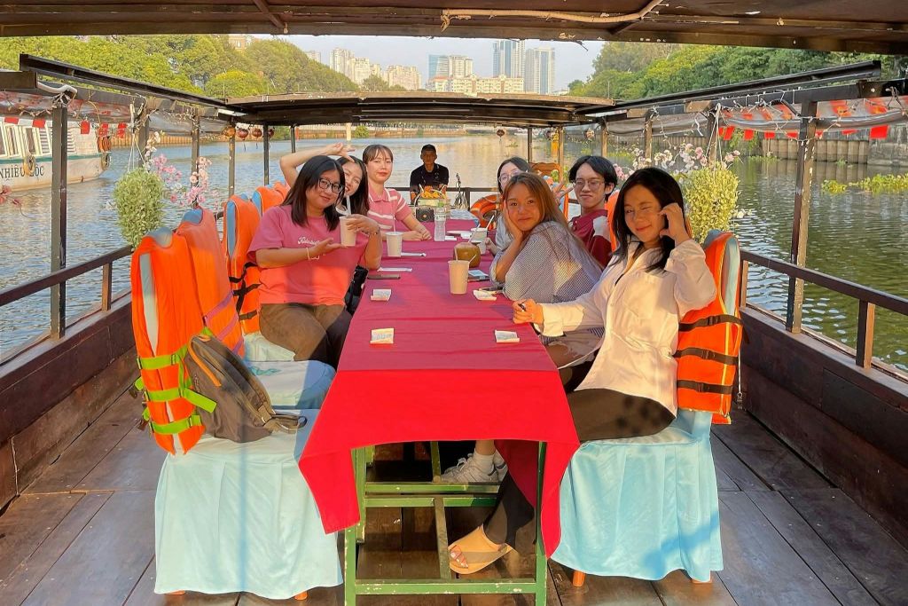 보트에 빨간 천이 놓인 테이블에 7명이 둘러앉아 물길과 도시 풍경을 배경으로 앉아 있습니다.