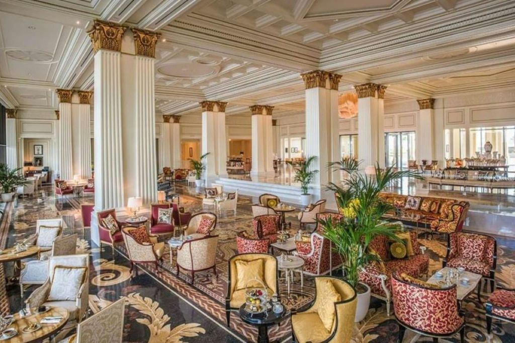 화려한 기둥, 무늬가 있는 카펫, 덮개를 씌운 의자, 화분, 샹들리에가 있는 우아한 호텔 라운지.