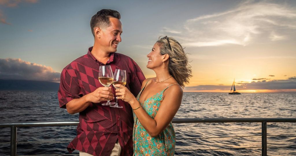 해 질 녘 보트에 선 두 사람이 와인잔을 들고 잔잔한 바다를 배경으로 미소를 짓고 있습니다.