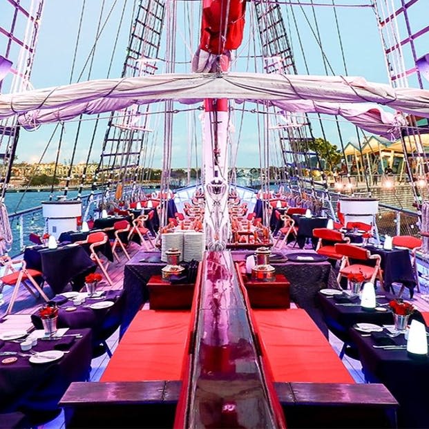 해질 무렵 항구 근처에서 빨간색과 검은색 테이블, 벤치, 오버헤드 리깅을 즐길 수 있는 식사용 선박 세트입니다.