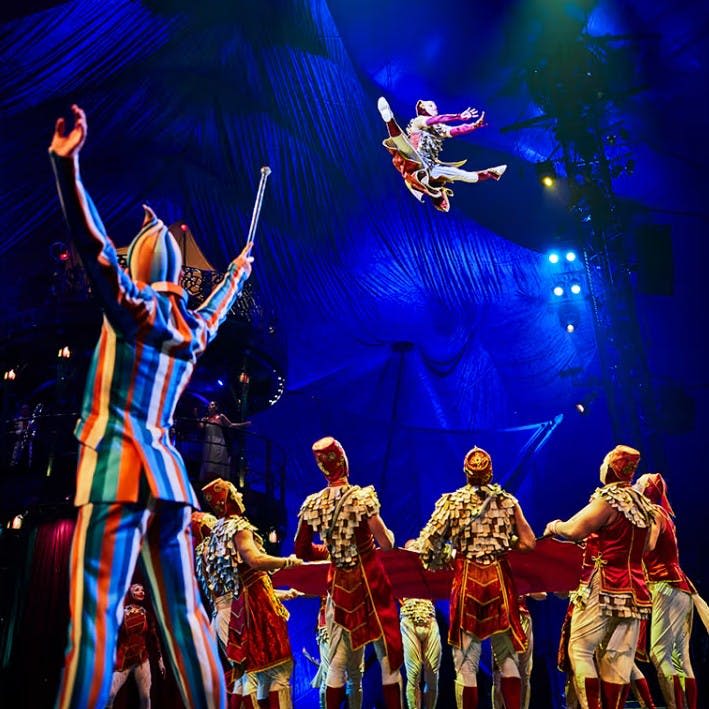 서커스 텐트에서 다채로운 의상을 입은 공연자들이 안전망을 들고 있는 그룹 위에 한 곡예사가 공중에서 매달려 있다.