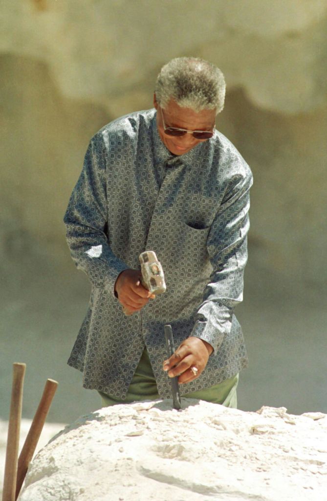 짧은 회색 머리카락과 선글라스를 가진 사람이 바위 망치를 들고 무늬가 있는 셔츠를 입고 흐릿한 모래 배경을 하고 있습니다.