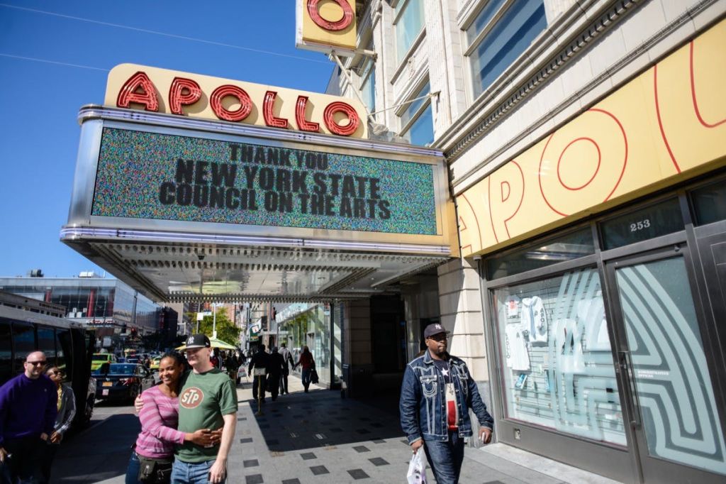 보행자들은 뉴욕주 예술위원회 (New York State Council on the Arts) 에 감사를 표하는 아폴로 극장 (Apollo Theater) 변두리 아래를 걷습니다.