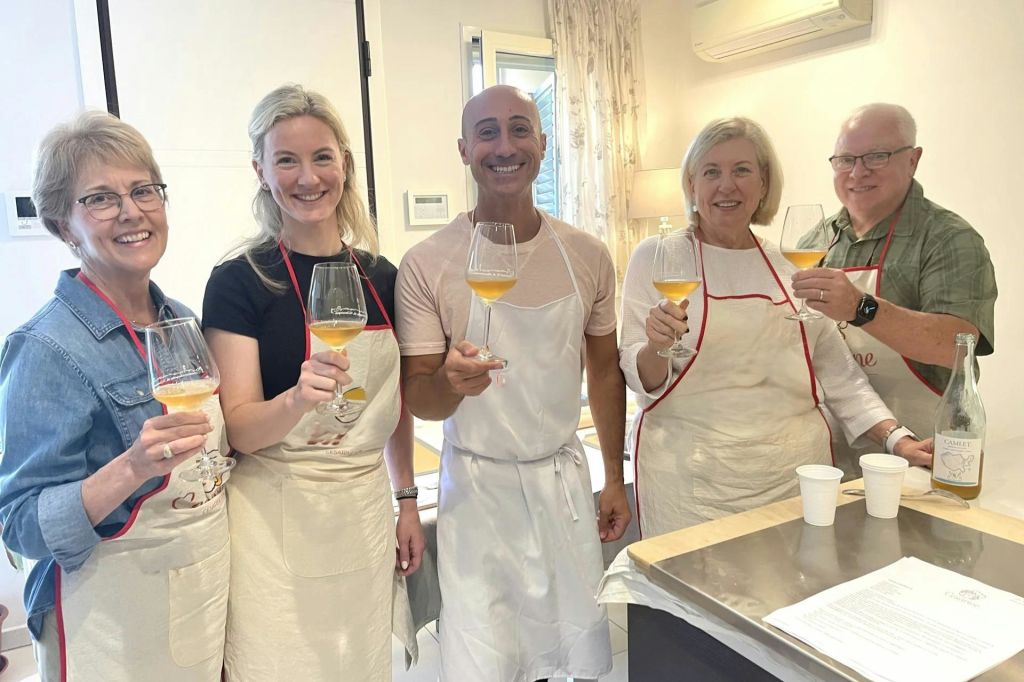 밝은 조명의 방에서 앞치마를 두른 세 사람이 오렌지 와인 잔을 들고 건배합니다.