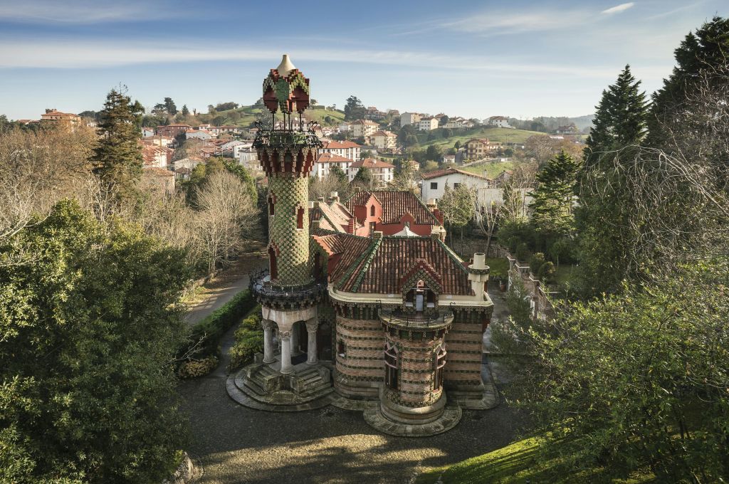높은 터렛이 있는 화려한 성 모양의 건물로, 나무로 둘러싸여 있으며 작은 마을과 언덕이 많은 지형을 배경으로 합니다.