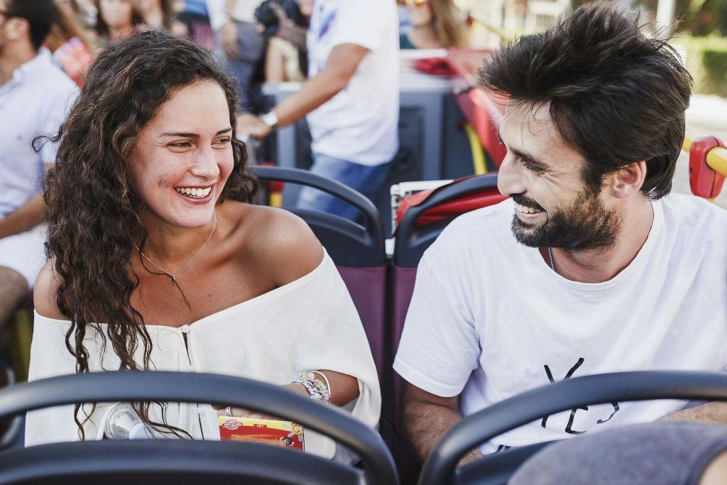 야외 투어 버스에 앉아 서로 웃고 있는 두 사람. 한 명은 간식 상자를 들고 있습니다. 배경에는 다른 승객들이 있습니다.