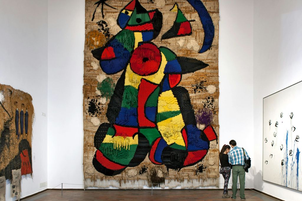 두 사람은 흰색 벽과 나무 바닥이 있는 아트 갤러리에서 크고 화려한 추상 태피스트리를 관찰합니다.