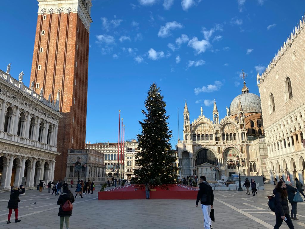 푸른 하늘 아래 종탑과 대성당을 포함한 역사적인 건물이 있는 광장에 커다란 장식의 크리스마스 트리가 서 있습니다.