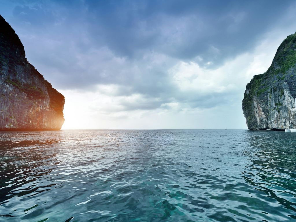 흐린 하늘 아래 양쪽에 우뚝 솟은 절벽이 있는 잔잔한 바다, 수평선을 엿보는 햇살.