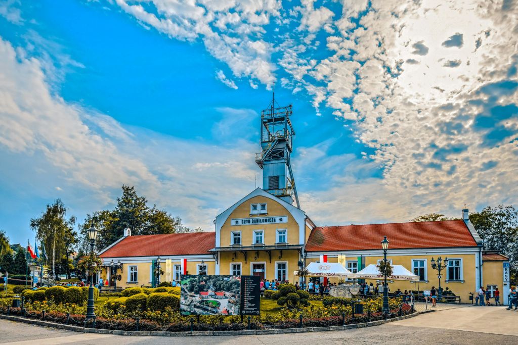 정원, 사람, 정보 표지판으로 둘러싸인 푸른 하늘 아래 시계탑이 있는 역사적인 건물.