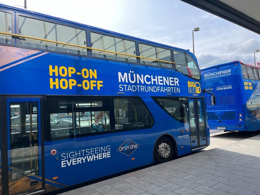 "Münchener Stadtrundfahrten" 과 "Hop-on Hop-off" 텍스트가 있는 파란색 2층 버스가 역에 주차되어 있습니다.