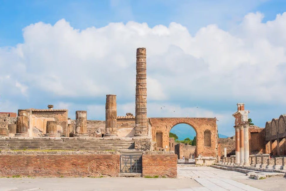 부분적으로 흐린 하늘 아래 기둥, 아치형 통로, 석조 구조물이 있는 고대 유적지.