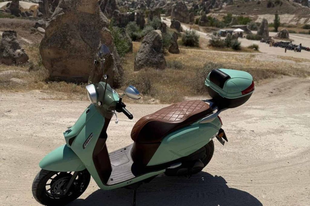 갈색 퀼트 시트와 후면 보관함이 있는 민트색 스쿠터는 바위 지형을 배경으로 먼지가 많은 길에 주차되어 있습니다.